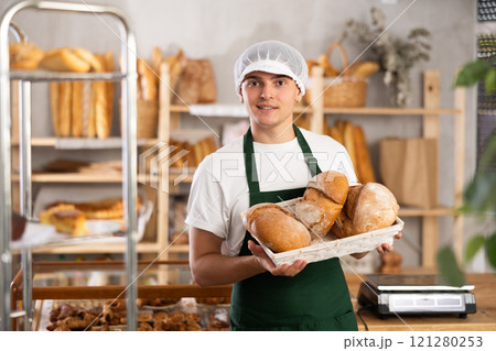 Young smiling man baker offers to buy fresh delicious round bread Young smiling man baker offers to buy fresh delicious round bread 121280253