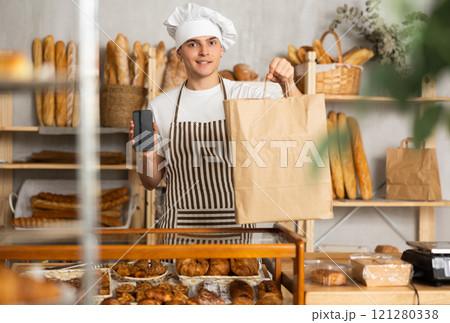 Guy seller in bakery hold out hand with phone, points at screen Guy seller in bakery hold out hand with phone, points at screen 121280338