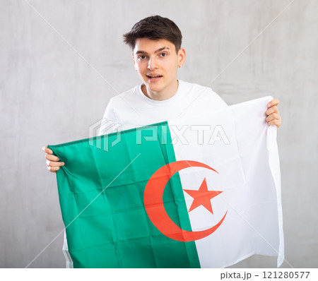 Reliant positive young man holds big national flag of Algeria with confident smile showing teeth. 121280577