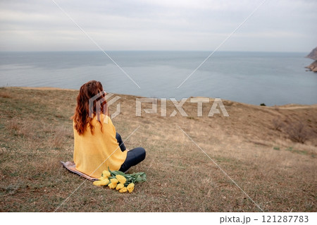 A woman sits on the grass with a bouquet of yellow flowers in her lap. She is looking out at the ocean, taking in the view. Concept of peace and tranquility. A woman sits on the grass with a bouquet of yellow flowers in her lap. She is looking out at the ocean, taking in the view. Concept of peace and tranquility. 121287783