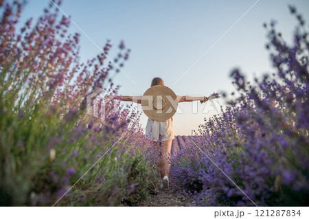 A woman is walking through a field of purple flowers with a straw hat on 121287834