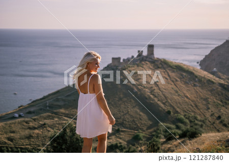 A blonde woman stands on a hill overlooking the ocean. She is wearing a white dress and she is enjoying the view. 121287840