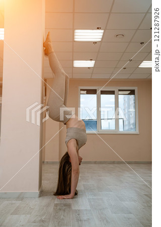 The girl is training in the yoga studio. Dressed in a beige tracksuit doing a handstand against the wall. Healthy lifestyle and yoga concept. The girl is training in the yoga studio. Dressed in a beige tracksuit doing a handstand against the wall. Healthy lifestyle and yoga concept. 121287926