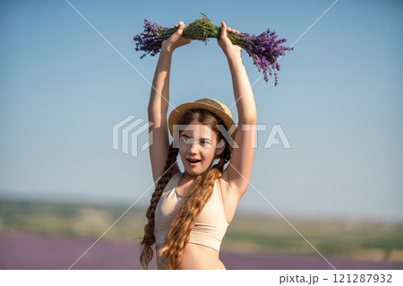girl is holding a bunch of lavender purple flowers in her hands and wearing a straw hat. She is smiling and she is enjoying herself. The scene is set in a field of lavender, which adds to the peaceful 121287932