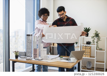 Indian man and African woman architects reviewing blueprint for modern city development. Creative office setting with desk models and laptop. Indian man and African woman architects reviewing blueprint for modern city development. Creative office setting with desk models and laptop. 121288826