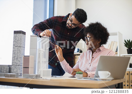 Indian man and African woman architects collaborate on model project for modern city. Diverse teamwork in bright office setting with creative discussion and urban planning focus. 121288835