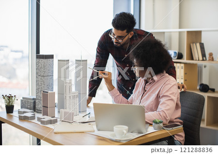 Indian man and African woman architects collaborate on model project for modern city. Diverse teamwork in bright office setting with creative discussion and urban planning focus. Indian man and African woman architects collaborate on model project for modern city. Diverse teamwork in bright office setting with creative discussion and urban planning focus. 121288836