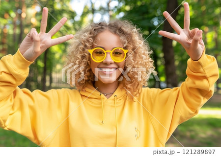 Portrait of a beautiful smiling curly blonde woman wearing yellow glasses and a sweatshirt outdoors in the park. Happy young woman showing peace symbol. People lifestyle concept. Copy space. Banner 121289897