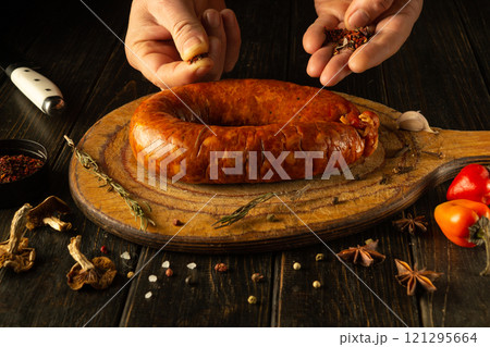 Hands skillfully season a smoked sausage on a wooden cutting board, surrounded by vibrant spices and fresh produce, creating an enticing culinary experience in the kitchen Hands skillfully season a smoked sausage on a wooden cutting board, surrounded by vibrant spices and fresh produce, creating an enticing culinary experience in the kitchen 121295664