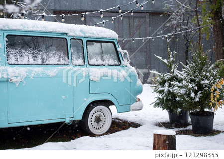 Retro blue van covered in snow, parked near pine trees and string lights in a snowy winter setting 121298355