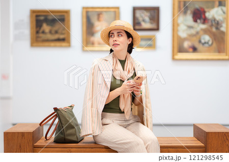 Mid shot of young Caucasian beautiful woman in an elegant suit is sitting on a bench in an art gallery and holds her smartphone. Modern cultural education in museum Mid shot of young Caucasian beautiful woman in an elegant suit is sitting on a bench in an art gallery and holds her smartphone. Modern cultural education in museum 121298545