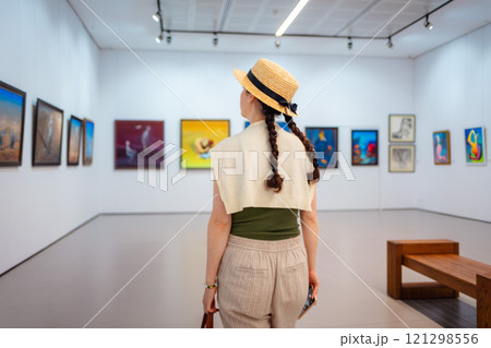 Back view of elegant woman in straw hat walking in the art gallery. Paintings in background. Concept of cultural education and Museum Day 121298556