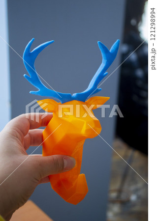 Person holding in hand an art object printed on 3D printer. Abstract toy model orange deer with blue horns created on 3d printer. FDM. Concept 3D Printing. Additive modern new progressive technologies Person holding in hand an art object printed on 3D printer. Abstract toy model orange deer with blue horns created on 3d printer. FDM. Concept 3D Printing. Additive modern new progressive technologies 121298994