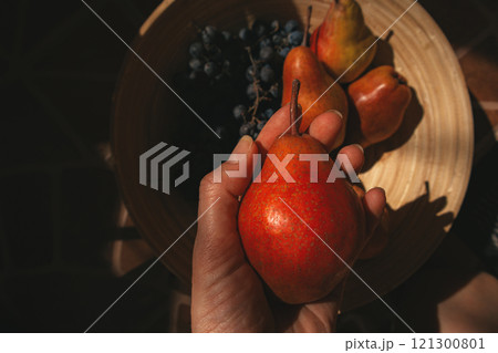 Ripe red pear in female hand view from above. Seasonal fruits - peaches, pears, blue grapes in a wooden round bowl, basket Rape organic summer harvest Ripe red pear in female hand view from above. Seasonal fruits - peaches, pears, blue grapes in a wooden round bowl, basket Rape organic summer harvest 121300801