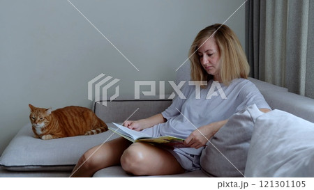 Young adult woman sits comfortably on couch absorbed in book while her cat lounges nearby. In living room caucasian female enjoys book with calm cat resting beside her reading and relaxation at home. Young adult woman sits comfortably on couch absorbed in book while her cat lounges nearby. In living room caucasian female enjoys book with calm cat resting beside her reading and relaxation at home. 121301105