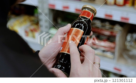 A customer in black studies the label on a bottle of sauce. Man carefully reads a product label in a busy supermarket aisle, making a healthy choice based on nutrition and ingredients A customer in black studies the label on a bottle of sauce. Man carefully reads a product label in a busy supermarket aisle, making a healthy choice based on nutrition and ingredients 121301112
