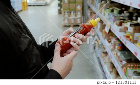 Shopper in a crowded supermarket aisle carefully examines a hot sauce bottle, trying to decide if it's worth purchasing among the variety of spices on the shelves 121301113