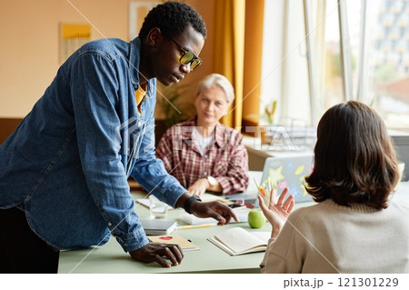 Side view of fashionable Black man in denim shirt outfit for office styled with colorful yellow glasses chatting with female colleague, while standing next to desk at workplace 121301229