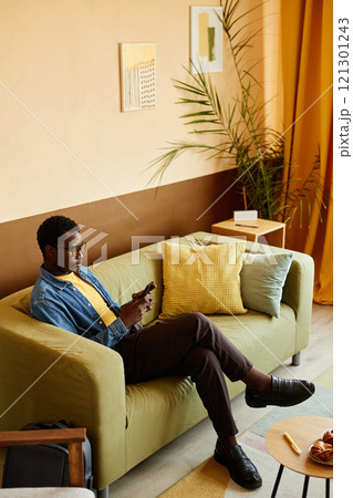 Vertical shot of young African American man scrolling through news feed on mobile phone, while sitting on comfy couch in cozy office featuring yellow interior accents 121301243