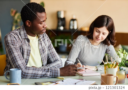 Medium shot of young African American man explaining details of business project to female coworker pointing at notes on paper, while sitting at meeting table in cozy office with beige walls Medium shot of young African American man explaining details of business project to female coworker pointing at notes on paper, while sitting at meeting table in cozy office with beige walls 121301324