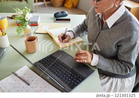 Cropped shot of self employed mature woman writing in notebook while using laptop at working desk composing business schedule at home office 121301325