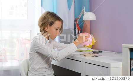 Young girl sits at her table in her room shaking coins from a pink piggy bank as she checks her savings. She explores the importance of personal finance and money management 121303879