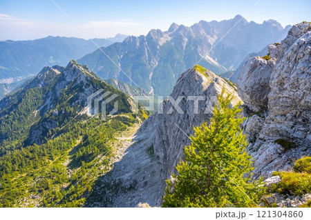 Clear skies reveal a breathtaking landscape of the Julian Alps in Slovenia. Rugged peaks rise dramatically, while lush greenery adorns the rocky slopes, showcasing nature's beauty. Clear skies reveal a breathtaking landscape of the Julian Alps in Slovenia. Rugged peaks rise dramatically, while lush greenery adorns the rocky slopes, showcasing nature's beauty. 121304860