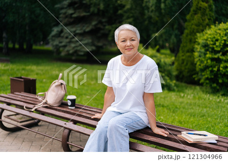 Thoughtful elderly woman with silver hair relaxing on park bench with backpack, books, and cup of coffee, savoring tranquility. Serene European gray-haired older female enjoying moment of relaxation. Thoughtful elderly woman with silver hair relaxing on park bench with backpack, books, and cup of coffee, savoring tranquility. Serene European gray-haired older female enjoying moment of relaxation. 121304966
