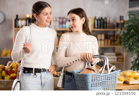 Two girls in shop choosing groceries and snacks, looking at something in windows, consulting, chatting 121309150