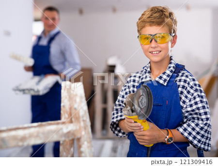 Portrait of little boy in protective glasses with an angle grinder in his hands in room being renovated Portrait of little boy in protective glasses with an angle grinder in his hands in room being renovated 121309416