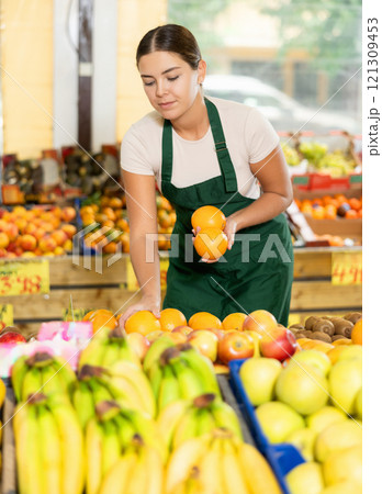 Interested salesgirl laying out oranges on display stand in supermarket Interested salesgirl laying out oranges on display stand in supermarket 121309453