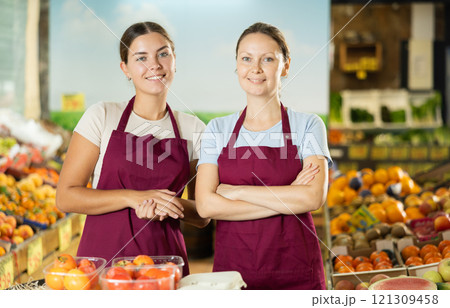 Friendly woman owner of fruit-vegetable shop standing in trading floor with girl assistant Friendly woman owner of fruit-vegetable shop standing in trading floor with girl assistant 121309458
