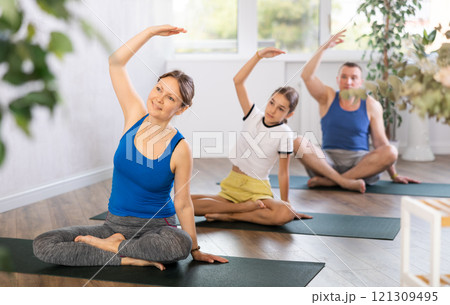 Middle-aged woman, man, preteen girl and boy warming up and doing side bends while sitting cross-legged before family Hatha yoga class in wellness studio 121309495