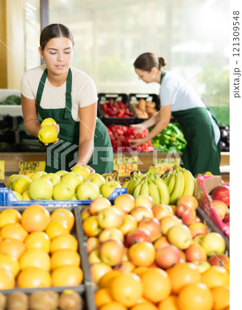 Girl seller of garden-stuff store works near showcase, sorting apples, checking goods Girl seller of garden-stuff store works near showcase, sorting apples, checking goods 121309548