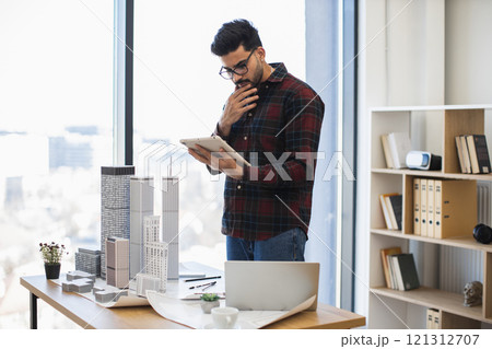 Indian male architect uses digital tablet to design modern city project. He stands in office near building models, displaying concentration and creativity. Indian male architect uses digital tablet to design modern city project. He stands in office near building models, displaying concentration and creativity. 121312707