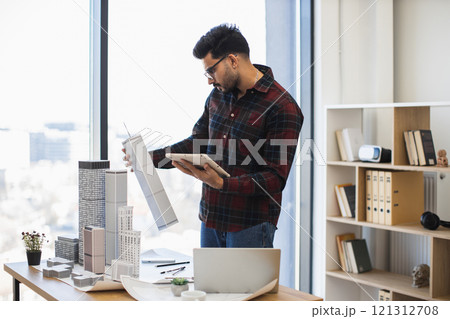 Young Indian male architect works on urban project using digital tools, creating city model in office. Focused on modern design and technology integration. Young Indian male architect works on urban project using digital tools, creating city model in office. Focused on modern design and technology integration. 121312708