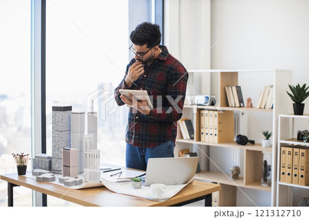 Indian male architect in modern office using digital tablet to develop city project with model buildings, emphasizing creativity and concentration. 121312710