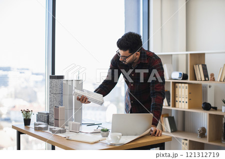 Indian male architect focuses on creating detailed city model for modern urban development, showcasing creativity and precision in office environment with natural light. Indian male architect focuses on creating detailed city model for modern urban development, showcasing creativity and precision in office environment with natural light. 121312719