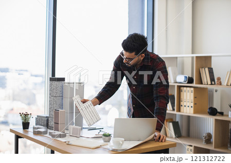 Indian male architect focuses on creating detailed city model for modern urban development, showcasing creativity and precision in office environment with natural light. 121312722