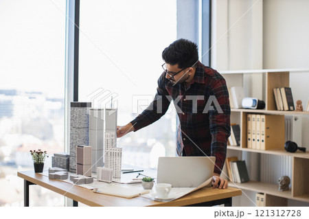 Indian male architect working on detailed model of modern city. Professional design concept showcasing creativity, planning, innovation. Bright office environment enhances focus and productivity. 121312728