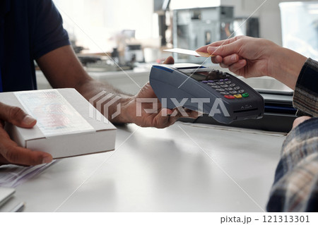 Close-up of hands during a payment transaction in a retail store, using a card reader. Person handing over card for payment, with boxes and store products in background 121313301