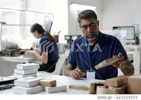 Bearded middle-aged man engrossed in examining packaging box at his desk in contemporary office setting with colleague working diligently in background 121313331