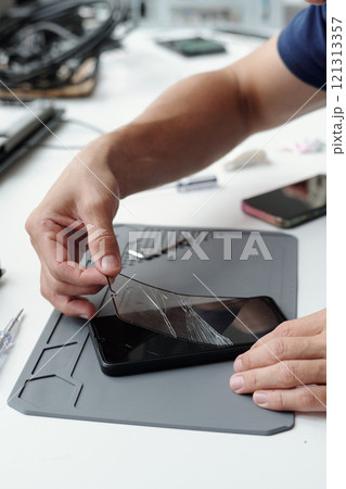 Close-up of hands repairing cracked smartphone screen using specialized tools on tech workbench. Many small components and electronic devices are scattered around Close-up of hands repairing cracked smartphone screen using specialized tools on tech workbench. Many small components and electronic devices are scattered around 121313357