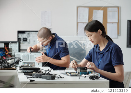 Two colleagues collaborating on electronics repair. Woman connecting hardware components while man working on other device. Office environment with various tools and devices present 121313359