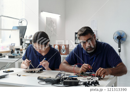 Two engineers working on electronic circuit boards, using tools and gadgets in a well-equipped lab. Both are focused on intricate details of their work 121313390