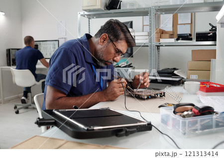 Concentrating man working on electronic circuit at modern office desk with computer and various tools in background. Another person is visible working at desk in background Concentrating man working on electronic circuit at modern office desk with computer and various tools in background. Another person is visible working at desk in background 121313414