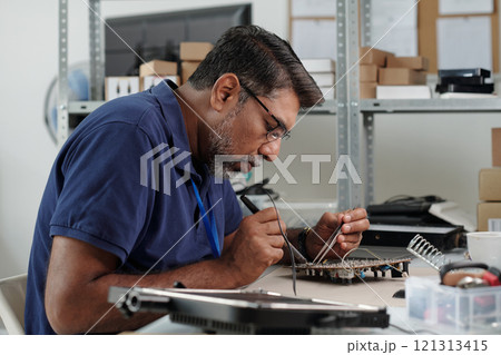 Man concentrating on fixing circuit board at workspace featuring shelves filled with boxes and equipment. Precision in handling small electronic components visible in workspace Man concentrating on fixing circuit board at workspace featuring shelves filled with boxes and equipment. Precision in handling small electronic components visible in workspace 121313415