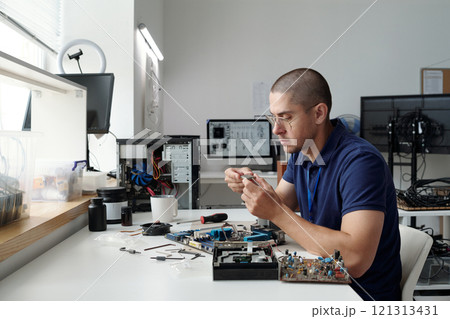 Man working diligently on electronic device at desk filled with components and tools. He is focused on intricate details while seated in well-organized workspace Man working diligently on electronic device at desk filled with components and tools. He is focused on intricate details while seated in well-organized workspace 121313431