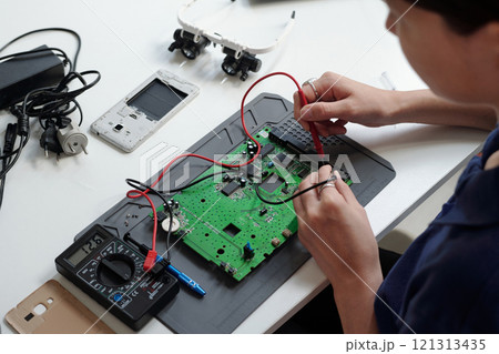 Detail of person working on electronic circuit board, using tools like multimeter and soldering iron. Various electronic components and devices are scattered on desk Detail of person working on electronic circuit board, using tools like multimeter and soldering iron. Various electronic components and devices are scattered on desk 121313435