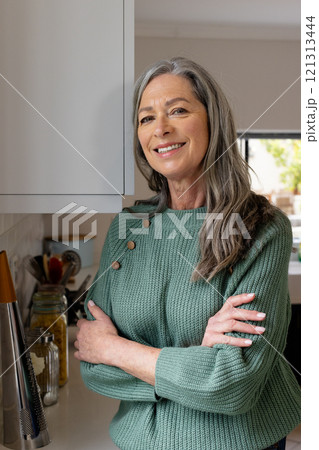 Smiling mature woman in cozy sweater standing confidently in kitchen, at home 121313444